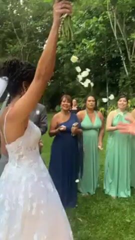 Here's a description of the video:

A bride in a white, floral lace gown with a long white veil over her dark, curly updo, is throwing her white floral bouquet to a group of women in green dresses. Confetti flutters in the air as the bouquet is tossed. The setting is an outdoor, possibly tropical, location with trees and foliage visible in the background. People are clapping and cheering. A photographer can be seen taking pictures to the side.