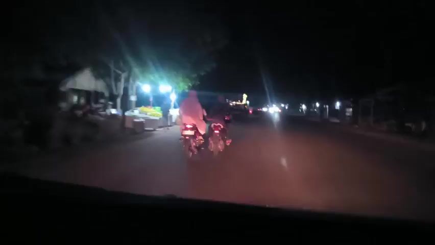 The video shows a night scene captured from inside a car, driving on a road illuminated by streetlights. Two motorcycles are driving ahead in the same direction, each carrying two people.  The person on the back of the first motorcycle is wearing a light-colored hijab or headscarf. The road is lined with small stalls or shops, some with people standing near them. Traffic is light, with a few other vehicles visible in the distance.
