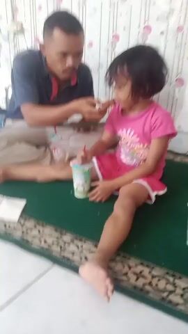 A man and a young girl are sitting on a green rug. The man is wearing a dark blue shirt and light-colored pants, and the girl is wearing a pink shirt and shorts. They both appear to be eating. There are two cups with straws on the rug between them, and a notebook is visible on the rug near the man. The background is out of focus but appears to be a wall with a patterned wallpaper. The lighting is dim.