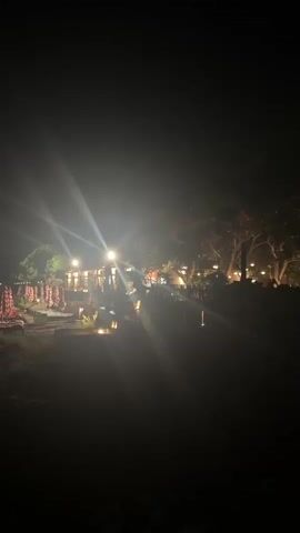 A nighttime video displays a dimly lit scene with strong lens flares from artificial lights. In the foreground, there is dark ground, possibly grass or soil. Further back, rows of closed beach umbrellas with red and white stripes are visible. Behind the umbrellas, a building with illuminated windows suggests a restaurant or resort. There are trees with scattered lights illuminating their branches in the background, and more trees are visible on the right side of the frame. The overall atmosphere is serene and possibly evokes a sense of an outdoor evening gathering or resort setting.