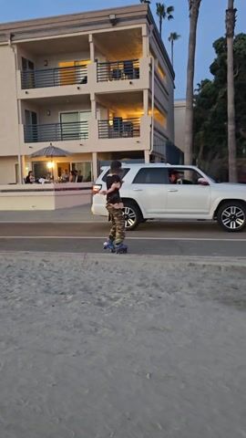 The video shows a person on roller skates standing on the edge of a beach, next to a paved walkway or road. The person is wearing a dark top and camouflage pants. Behind them is a three-story beige building with balconies and some people sitting under an umbrella at a table. A white SUV is driving past on the road. Palm trees are visible in the background. The scene appears to be at dusk or in the early evening. The sky is a mix of blue and orange.