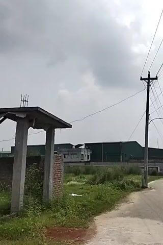 The video depicts a rural or industrial area on an overcast day. The camera is moving, presumably mounted on a vehicle, and captures views of buildings, fields, and infrastructure. Predominantly, the scenery includes a long, dark-colored industrial building, utility poles with numerous power lines, and grassy fields. There's a blue, two-story building with what appears to be an unfinished roof. The sky is cloudy, hinting at potentially inclement weather. The road is a mix of paved and unpaved sections.