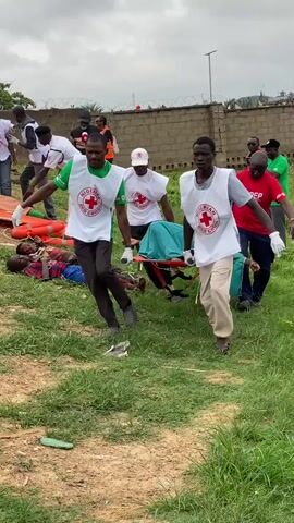 The video shows a group of people in a grassy, outdoor area. Several individuals wearing red cross vests are carrying stretchers with what appear to be people on them. They are walking across the uneven terrain. In the background, there are more people, some wearing red and white clothing. There is a building or wall in the distance. The sky is overcast. The overall impression is one of a rescue or medical assistance operation.