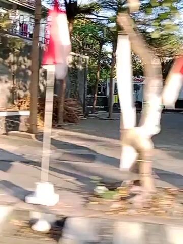 The video shows a street scene from a moving vehicle. In the foreground, there is a blurry brown object, possibly wood debris. Beyond that, a grey asphalt road is visible, with a white and yellow striped curb on the right side. On the left side of the road, a white wall with brickwork at the base is partially visible, with some stacked wooden pallets behind it. Further down the road, a yellow truck is driving away, showing its side and back. The truck has a black tarp covering its cargo area and a driver visible in the cab. To the right of the truck, there is a building with a blue, collapsible gate. A flag pole with a red and white flag is also visible next to the gate. Continuing down the road, a white building with multiple windows and balconies is seen. A red metal railing with yellow poles runs along the front of a property with a stone wall at its base. Finally, a silver car is driving past on the road. The sky is clear and appears to be daytime.