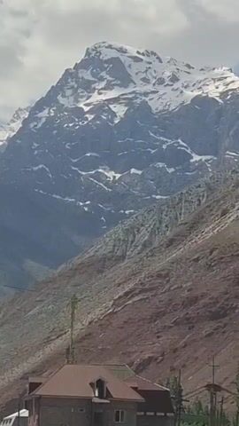 The video shows a snow-capped mountain peak framed by a cloudy sky. A brown building is seen at the base of the mountain, and the camera moves upward, revealing the back of a large orange truck. The focus shifts to the mountain again, emphasizing its snow-covered peak against the sky. The truck intermittently obstructs the view of the mountain. There is musical background noise, implying the presence of people near the camera but they are not visible.