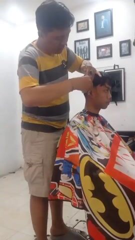 A male barber is cutting the hair of a male client. The barber is wearing a gray, yellow, and black striped t-shirt and beige cargo shorts. The client is wearing a colorful cape featuring Batman imagery. The barber is using scissors and a comb to cut the client's hair. The background appears to be a room with framed pictures on the wall. The lighting is bright, suggesting an indoor setting.