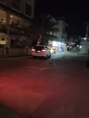 The video shows a street scene at night. A white SUV is parked on the right side of the street. The SUV's brake lights are illuminated red. There are buildings on the left and right sides of the street, some with lights on. A palm tree is visible behind the SUV. The street is paved and appears to be wet in places, reflecting the red light from the brake lights. There are some parked motorcycles in front of a building on the right.