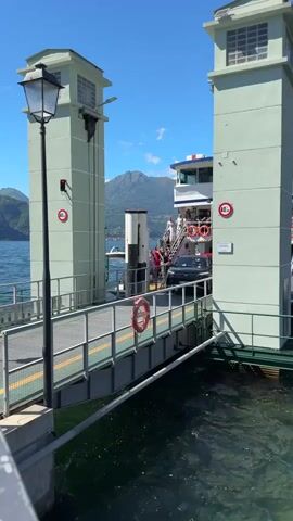 A car is being driven onto a ferry. The car is a dark colored sedan. The ferry appears to be docked at a terminal with two large green towers on either side of the ramp. There are people on the ferry, some standing on stairs. The ferry is on a body of water, likely a lake, with mountains visible in the background. The sky is clear and blue. The time of day appears to be daytime, with bright sunlight.