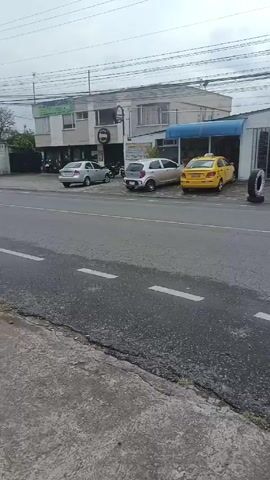 The video depicts a street scene during the daytime. The camera is positioned on a sidewalk, facing a road with white lane markings. In the initial frame, a tire shop called "Torres Tires" is visible on the right. A yellow taxi is parked near the shop. As the video progresses, the camera pans from left to right, showcasing various commercial establishments and parked cars lining the street. A green dumpster sits on the sidewalk, and a bare tree and some shrubbery are seen behind it. Overhead, power lines stretch across the sky, which is covered with clouds. The scene appears relatively quiet, with only a few vehicles visible on the road.