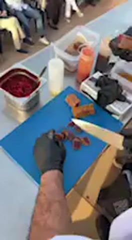 The video captures an outdoor culinary demonstration in Barbate, Spain, focusing on a person in black gloves meticulously slicing reddish-brown food on a blue cutting board with a chef's knife. Surrounding the cutting area on a light-colored table are various culinary items, including a metal container holding chopped red ingredients, multiple squeeze bottles containing different liquids (red, white, and brown), and a white plate with a dollop of red sauce. In the background, an audience is seated, observing the ongoing food preparation. The setting appears to be a daytime event with bright, natural lighting. The activity is centered around precise food preparation, likely a cooking demonstration or part of a food festival. No visible text is present in the frame. The audio features spoken Spanish, where culinary terms like "saborizante" (flavoring) and "caldo dashi" (dashi broth) are discernible, indicating a discussion about the food being prepared or its ingredients.