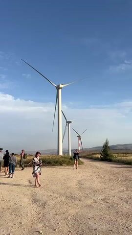 The video shows a group of people near three wind turbines in a rural, dry, and somewhat dusty area. The wind turbines are tall and white, with one having red and white stripes near the top. The sky is blue with some scattered clouds. A group of people are standing near a few parked cars, some are looking in the back of an SUV. A woman in a short dress or top with straps is walking towards the camera in the foreground. The environment appears to be a field or open space with low-lying vegetation.