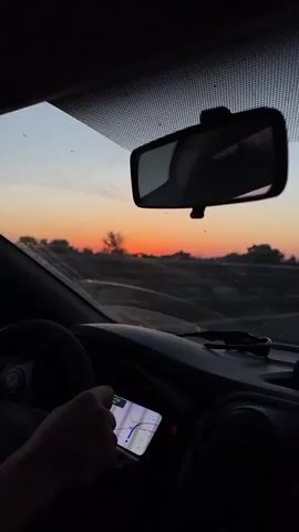A first-person perspective shot from inside a car shows a hand holding a smartphone with a navigation app displayed on the screen. The phone is positioned on the dashboard in front of the driver. The view through the windshield reveals a sunset with a vibrant orange and pink sky. Trees and a road are visible in the distance. The rearview mirror is also in frame, reflecting the sunset. The overall mood is calm and suggests travel during the golden hour.