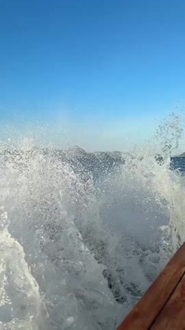 The video shows a view from a boat moving quickly on the ocean. The camera is pointed toward the rear of the boat, showing the wake created by the boat cutting through the water, which is a frothy white. The water is a deep blue, and in the distance there are small islands with dark, rocky terrain and a few green patches. The sky is a clear, bright blue. The boat appears to be made of wood.