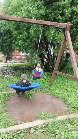 Two young children are playing on a wooden swing set in a grassy yard. One child, wearing a dark shirt and pants, sits on a blue disc-shaped swing. The other child, wearing a brightly colored jacket and gray pants, is standing and pushing off the ground, appearing to get momentum to swing. The ground beneath the swings is wet and muddy. Trees and a brick building are visible in the background.