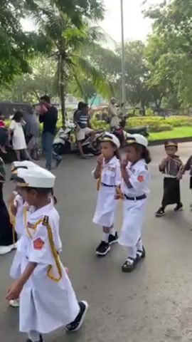 A group of young children dressed in various uniforms are walking in a park. Two girls in white sailor uniforms are in the front. Behind them are several boys in police and military style uniforms. They are walking on a paved path. In the background, there are trees, grass, and parked motorcycles. Several adults are standing nearby, observing the children.