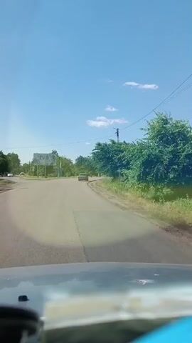 The video shows a first-person view from a vehicle driving on a paved road. The sky is clear and blue with some scattered clouds. Trees and vegetation line the sides of the road, and in the distance, there is a billboard with a colorful image. There are a few other vehicles visible on the road, and some puddles of water on the ground. The overall impression is of a sunny day in a rural or suburban area.