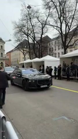 The video shows a motorcade of black vehicles driving down a street in an urban setting. The first car appears to be a BMW, followed by several other black cars, including what appears to be an Audi SUV. People are standing on the sidewalks and near white tents set up on the side of the road. Some of the people are dressed in suits. The buildings are tall and appear to be located in Europe. The sky is overcast. Yellow and blue flags are visible on a building, suggesting support for Ukraine. A man in a suit is walking in front of the cars.