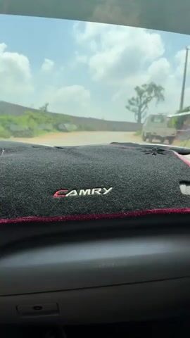 A static shot from inside a car, likely a Toyota Camry, looking out the windshield at a road ahead. The car's dashboard is visible, covered with a black mat embroidered with "CAMRY" in red. A car radio and what seems to be a device like a parking aid are on the dashboard. Through the windshield, there is a road with a brick wall on the left and a tree on the right. A white truck is also visible on the right. The sky is partly cloudy. The car is stationary.