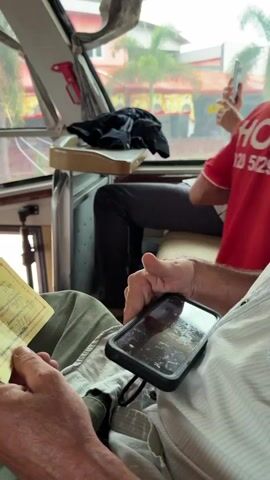 The video shows a close-up view of two individuals inside a vehicle, likely a bus, based on the seating and the windows. In the foreground, a person's hands are visible holding a smartphone and a yellow ticket. The phone's screen displays a reflection of the ticket. The person is wearing a light-colored, possibly checkered shirt and green pants. Their fingers are touching the phone screen, suggesting they are interacting with it. To the left of the main subject, another yellow ticket is partially visible, held by a hand wearing a green garment. In the background, another person is seated, holding a white object, possibly a phone, and a yellow cup. This individual is wearing a light-colored shirt and dark pants. The interior of the vehicle appears to have beige seating and a beige tray table. Through the windows, a blurred outdoor scene is visible, with buildings and greenery, indicating movement. The lighting suggests it is daytime.