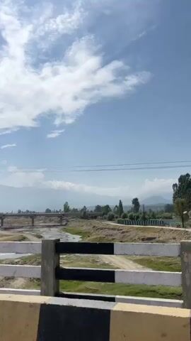 A video shows a wide view of a landscape. In the foreground, a white barrier with black and gray stripes is visible, suggesting it might be part of a bridge or roadside. Beyond the barrier, a dry riverbed with a small stream is present, spanned by a concrete bridge. In the distance, mountains form the backdrop under a partly cloudy sky. Trees are scattered throughout the scene, and a dirt road winds its way through the landscape. The overall impression is of a rural or semi-rural environment.