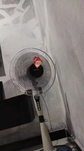 A person's legs and feet are visible at the bottom of the frame. A pedestal fan is standing on the floor in the center of the frame. The fan has a wire cage, and a tag is attached to the front of the fan. The fan is positioned near a dark rectangular object. The background is a wall with a rough texture.