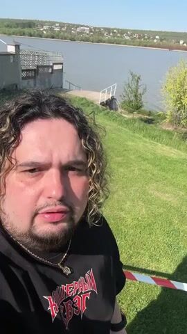 The video features a man standing outdoors, facing the camera. He is wearing a black t-shirt with a red graphic on it, and a gold chain around his neck. He has curly hair. Behind him is a green lawn, and in the distance, there is a lake. As the video progresses, the camera pans to reveal a stage setup further into the lawn area. There are trees in the background, and the sky is blue.