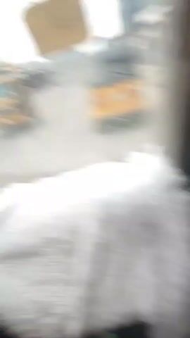 The video captures a street scene from inside a vehicle. The foreground is dominated by a fluffy white material, likely a seat cover or blanket. Through the windshield, a dusty, paved road is visible with some debris. On the right side of the frame, a yellow auto-rickshaw is driving away from the camera. Further down the road, there are buildings on both sides, some appearing to be shops or homes. People are walking on the sidewalk and in the road. The sky is overcast. Two yellow rectangular stickers are affixed to the windshield, partially obstructing the view. One sticker has text, possibly a date. The camera pans slightly, offering glimpses of the side view mirror and the side of the vehicle.