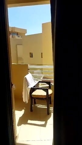 The video opens with a view of a balcony from inside a room, looking out through an open doorway framed by dark curtains. The balcony is furnished with a wicker chair with a light-colored cushion. A white towel is draped over the back of the chair. A book is also visible on the chair. The balcony floor is tiled. Beyond the balcony railing, there are two-story buildings with yellow facades and windows. The sky is clear and blue. The camera pans slightly to the right, revealing more of the buildings and the surrounding architecture, which appears to be a resort or hotel complex. The sunlight casts shadows on the balcony. The video ends with a view of the buildings stretching into the distance.