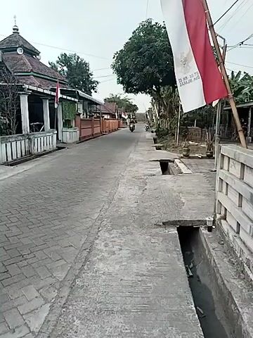A paved road with brick sidewalks lines the left side of the frame, with a drainage ditch on the right. Several Indonesian flags, red and white, are visible along the road, one prominently displayed on the right, and others in the distance. A building resembling a mosque with a dome is on the left side of the road, and other buildings are visible further down. A person on a scooter rides down the road towards the viewer, and another person is visible further in the distance. The sky is overcast.