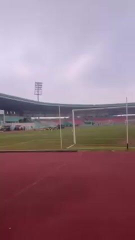 The video shows a group of people at a stadium with a running track. The stadium has a covered seating area and a large screen. People are stretching and walking on the track and the surrounding grassy areas. The video focuses on the track and grassy area, showing the texture of the ground. A person is seen walking on the grass.