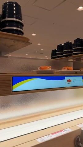 The video shows a conveyor belt sushi restaurant. Several plates of salmon sushi are visible on the belt, moving past a digital display that shows a picture of salmon sushi and indicates "Salmon 4 dish". In the background, stacks of dark colored bowls are visible. The lighting appears to be from overhead fluorescent lights.