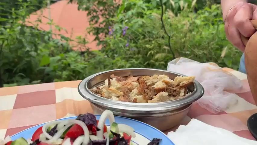 A person's hands are holding a rustic, brown, clay pot and tilting it to pour a yellow, soupy liquid containing pieces of meat and potatoes into a metal bowl filled with bread cubes. The bowl is sitting on a checkered tablecloth. In the background, lush green foliage and a glimpse of a red-tiled roof are visible. A blue plate with salad is partially visible in the foreground. A piece of white mesh fabric is draped over the side of the pot.