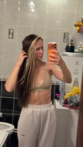 In a bathroom with tiled walls and floor, a woman stands facing a mirror, holding an orange smartphone in her hand, presumably taking a selfie. She is wearing a beige sports bra and light gray sweatpants. Her hair is long, dark brown with a prominent blonde streak near the front. The hair appears wet. The bathroom contains a toilet with a cartoon face sticker on the lid, a washing machine, a water heater, and various toiletries and cleaning supplies are visible on the counter and shelves. The woman turns her head and flips her hair as she takes the photo.