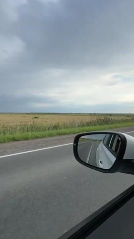 The video is a vertical shot from inside a car, looking out the side window and into the side-view mirror. The car is on a paved road, which runs horizontally across the lower half of the frame. To the left of the road is a field of tall, dry grass or wheat, stretching to the horizon. Beyond the field, the sky is filled with heavy, gray clouds, suggesting overcast weather. In the side-view mirror, the back of a white car is visible, also traveling on the same road. The reflection in the mirror shows the road ahead and a hint of the landscape. The interior of the car, including a portion of the door and window frame, is visible in the foreground on the right side of the frame. The overall scene depicts a journey through a rural landscape under a cloudy sky.