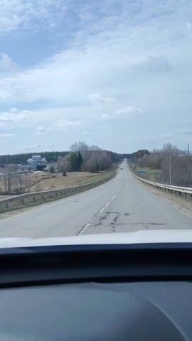 The video captures a first-person perspective from a vehicle traveling on a two-lane paved road. The time of day is daylight, with a partly cloudy sky showing patches of blue. The weather appears fair.

The road ahead stretches into the distance, with a slight incline. It has a solid white line on the right shoulder and a dashed white line separating the two directions of traffic. The asphalt shows signs of wear, with visible cracks and patches. Metal guardrails line both sides of the road.

To the left of the road, there are some buildings, possibly houses, nestled among mostly bare trees and fields. The right side of the road also features fields and trees, many of which are deciduous and without leaves, suggesting late winter or early spring.

Several cars are seen driving in the opposite direction on the road.

At the 0:02 mark, a blue road sign with white text becomes visible on the right side of the road. The text clearly reads "р КАЗМАСКА," which likely refers to "river Kazmaska" or a settlement named Kazmaska. This detail aligns with the provided location context of Novaya Kazmaska, Russia. The scene depicts a routine drive through a rural or semi-rural landscape.