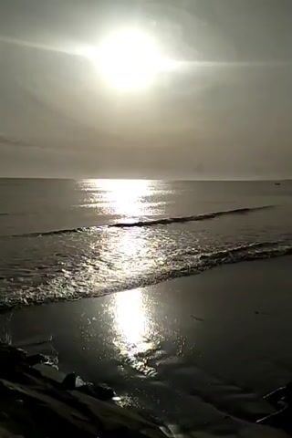 The video shows a body of water with waves gently lapping onto a dark sandy beach. The sun is high in the sky, casting a bright glare on the water's surface, creating a shimmering path of light. The sky is overcast, with the sun appearing as a bright, undefined orb. The foreground shows a dark, rocky shore with some vegetation. The scene appears to be a natural landscape, likely a coastline at sunrise or sunset, given the low angle of the sun and the soft light. There are no people or man-made objects clearly visible in the frame.