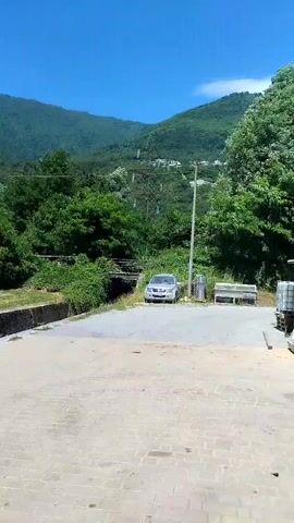 The video captures an outdoor scene with a clear blue sky and green, rolling hills in the background. In the foreground, there is a paved area and a concrete wall. A metal excavator bucket rests on the paved surface. Further back, a silver car is parked near some trees and a power pole. The landscape appears to be a rural or semi-rural area, possibly a construction site or a place undergoing some work, given the presence of the excavator bucket.