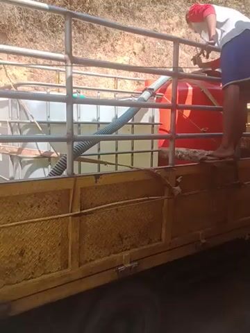 A person is seen operating a water truck with a large white water tank. The person is leaning over and appears to be adjusting something near the top of the tank. The truck has a yellow cargo bed with a metal cage structure. The environment appears to be dry and outdoors, possibly a rural area with sparse vegetation.