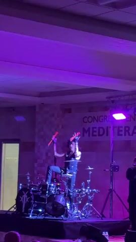 A male drummer, dressed in black with long dark hair, performs on a drum set on a stage illuminated by pink lights. He is seated and is actively playing the drums with drumsticks. The backdrop features the words "CONGRESS CENTER MEDIT" in white lettering. There is a person on the far right of the frame, partially visible.