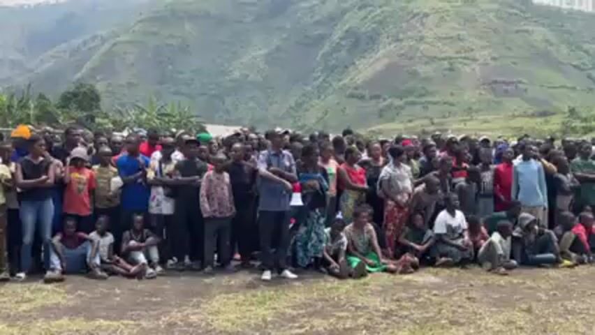 A medium shot shows a large group of people gathered in a grassy field. Most are standing while some sit on the ground. A man in the center of the group holds a megaphone. Behind the crowd, there is a wide riverbed and a mountain range. The sky is overcast.
