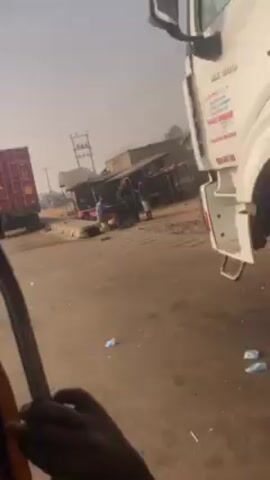 The video shows a roadside scene, likely in a developing country. A red shipping container on a large truck is moving along a paved road. Alongside the road are several makeshift stalls, appearing to be small shops. People are visible near these stalls, going about their business. The environment has a dusty or hazy atmosphere. The perspective is from a vehicle, possibly a smaller motorcycle-style vehicle, moving alongside the truck.