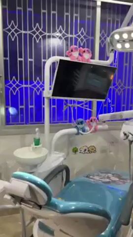 A person is recording video inside a dentist's office. The dentist's chair is turquoise and decorated with cartoon characters. The dentist's equipment, including a light and a monitor, is in place. Plush dolls are attached to the equipment. Behind the dentist's chair is a window with blue lighting. The person recording is wearing a black jacket.