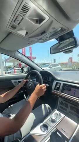 The video shows the interior of a car from the passenger's perspective, looking forward. A person's hands are visible on the steering wheel, indicating they are driving. The dashboard features a navigation screen displaying a map. Outside, the scene is bright with a clear blue sky and some trees. A gas station with red pumps is visible on the left. The car appears to be moving on a road with other vehicles and buildings in the distance. In one shot, a hand is raised, obscuring the view momentarily. The interior of the car has wood trim accents on the dashboard and center console.