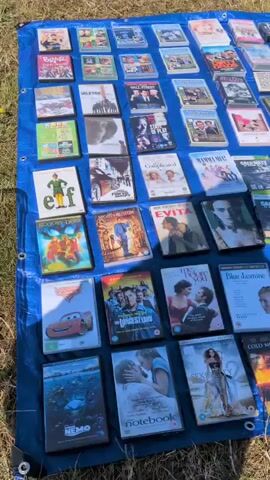 The video shows a close-up aerial view of a collection of DVDs and video games displayed on a blue tarp spread on the ground. The items are arranged in rows, and the camera pans across them. The collection appears to be for sale at an outdoor market or yard sale. Some of the titles visible include "Cars," "The Longest Yard," "The Notebook," "Mamma Mia!," "Evita," "The Duchess," and various "Call of Duty" and "Assassin's Creed" video games. The lighting suggests it is daytime, and the ground beneath the tarp is covered in dried grass or dirt, indicating an outdoor setting. There are no people or animals clearly visible in the frame.