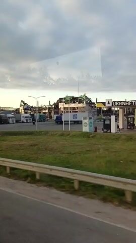 The video shows a parking lot with several semi-trailer trucks parked. In the background, a large building is heavily damaged, with its roof partially collapsed and exposed framework. Construction equipment, possibly for demolition or cleanup, is visible near the damaged building. The sky is overcast with clouds. The scene appears to be during daylight hours.