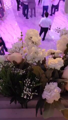 The video captures a bustling indoor event, likely a wedding reception or formal party, viewed from a high vantage point. The foreground is dominated by a large floral arrangement of white and green flowers. The midground shows a dance floor where several men in white shirts and dark pants are dancing in a circle, some with arms around each other. Other guests are seated at tables arranged around the perimeter of the room. The room itself is elaborately decorated with cascading white crystal chandeliers and floral accents. The lighting is dramatic, with pink and yellow spotlights illuminating the dance floor. In the background, more guests are visible, and the overall atmosphere appears festive. The flooring is a light-colored marble with reflective qualities. The camera appears to be positioned on a balcony or elevated platform overlooking the main event space.
