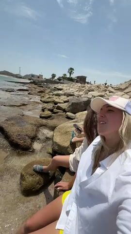 The video captures two women enjoying a sunny day on a rocky coastline in Dhadna, United Arab Emirates. One woman with blonde hair, wearing a white shirt, yellow shorts, and a white cap, is seen sitting on a large rock with her feet near the water. Her companion, with long dark hair, also in a white shirt, sits on another rock nearby, holding a phone.

The scene transitions to a wide view of the calm, clear blue-green sea, showing the rocky shoreline extending into the distance. Gentle waves lap against the rocks. The horizon features distant land formations and what appear to be coastal structures. The sky is bright blue with scattered white clouds, indicating clear and pleasant weather. Palm trees are visible in the background, adding to the tropical feel of the location. The video then pans back to the women, with the blonde-haired woman seemingly talking or singing with an animated expression. The overall impression is one of relaxation and leisure by the sea.