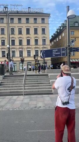 This media file captures a skateboarding session in Stockholm, Sweden. The main subject is a young man on a skateboard performing a trick down a set of stairs. He is wearing a black and white striped t-shirt, black pants, a dark beanie, and white sneakers. He successfully lands the trick, with another person in the foreground filming him with a camera. This person is wearing a white t-shirt, red pants, and a beige hat, with a camera hanging from their shoulder.

The setting is an urban environment with a large, light-colored building in the background featuring multiple windows and ornate detailing. Above the building, a sign reads "DAGENS NYHETER" in red letters. Another signpost points towards "VÄLLINGBY," "SOLNA," and "Centralstation City," indicating a city location. There are traffic lights, a pedestrian crossing, and other people in the background, suggesting a public square or street. The weather appears to be clear with a partly cloudy sky. The time of day seems to be late afternoon or early evening, given the lighting conditions. The overall mood is energetic and focused on the skateboarding activity.