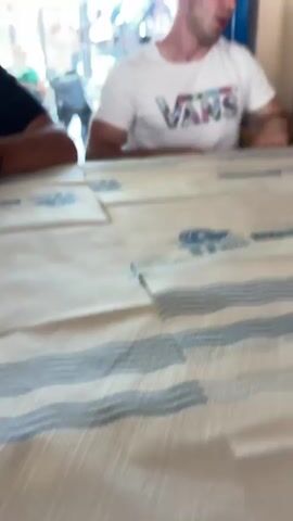 Two young men are sitting at a table in a restaurant. The table has a paper tablecloth with blue designs. One man is wearing a black shirt and gives a thumbs up while holding a phone. The other man is wearing a white Vans t-shirt. The camera pans around the restaurant, showing empty tables and chairs, a drink refrigerator, and people walking outside. A third young man wearing a tan "Flair" t-shirt and matching shorts walks past the camera.