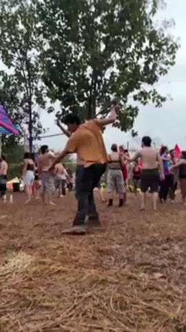 The video shows a crowd of people at an outdoor event on what appears to be a muddy ground covered in hay. Many are shirtless, dancing or moving to music, and some are partially clothed. There is a person in the foreground wearing a yellow shirt and dark pants who is actively dancing and gesticulating with a stick-like object. The surroundings include trees and decorated structures with colorful flags, indicating a festival or outdoor gathering.